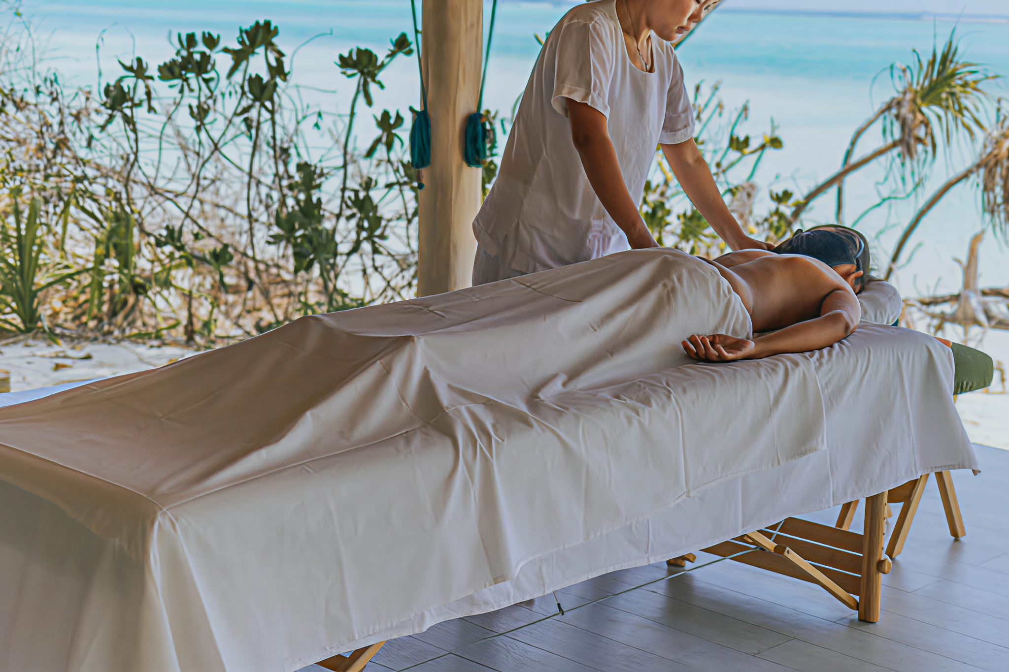 Spa therapist performing a massage on a guest in an open-air treatment area with ocean views framed by tropical vegetation.