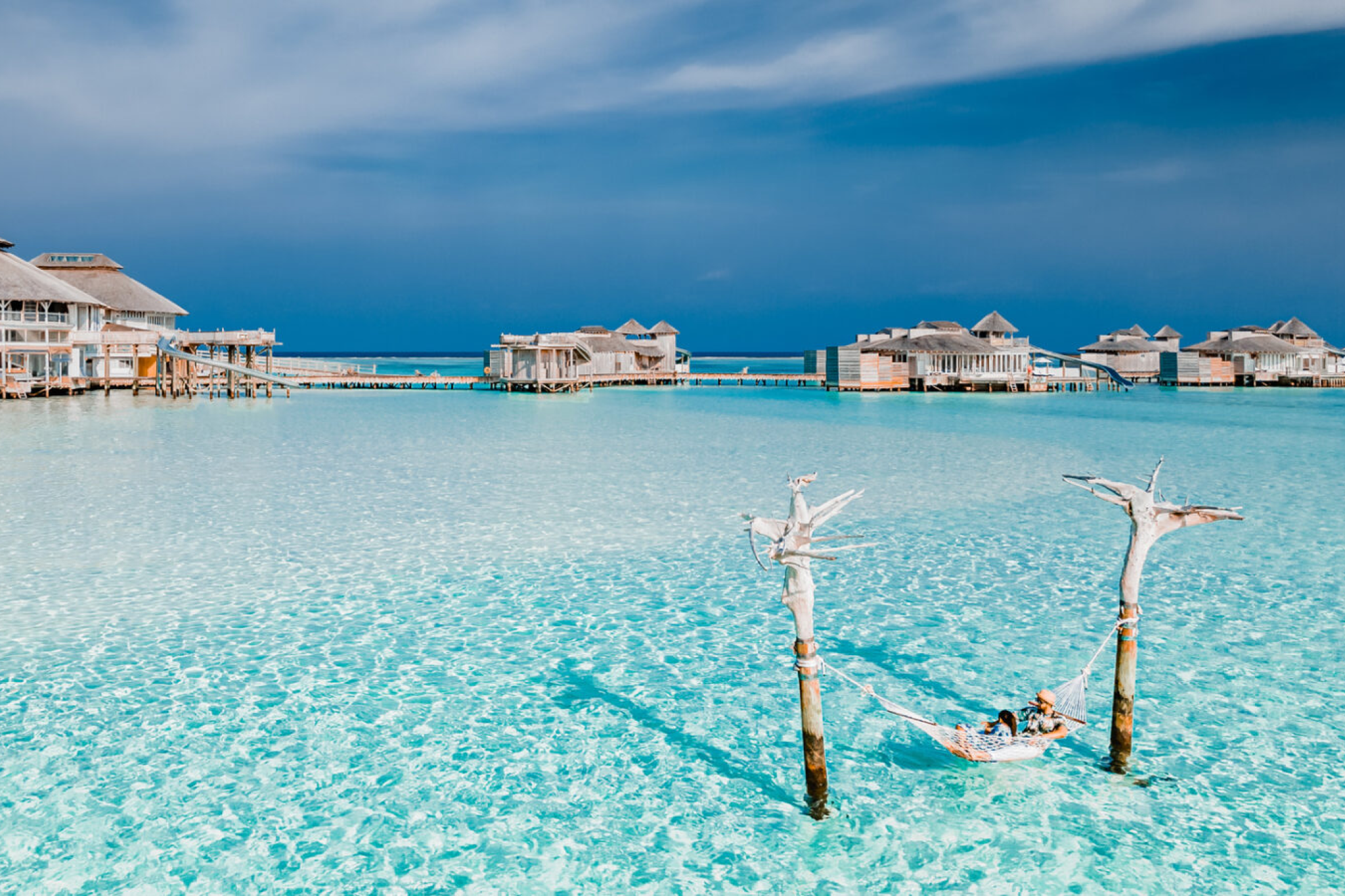 Couple relaxing in a hammock suspended over shallow crystal-clear waters, with overwater villas of Soneva Jani in the background.