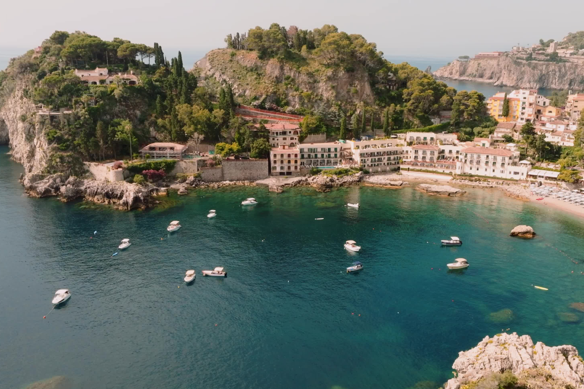 A stunning aerial perspective of Belmond Villa Sant’Andrea nestled along Taormina’s turquoise bay, with anchored boats dotting the shoreline—an idyllic coastal haven featured as one of the top beach resorts in Europe in 2025.