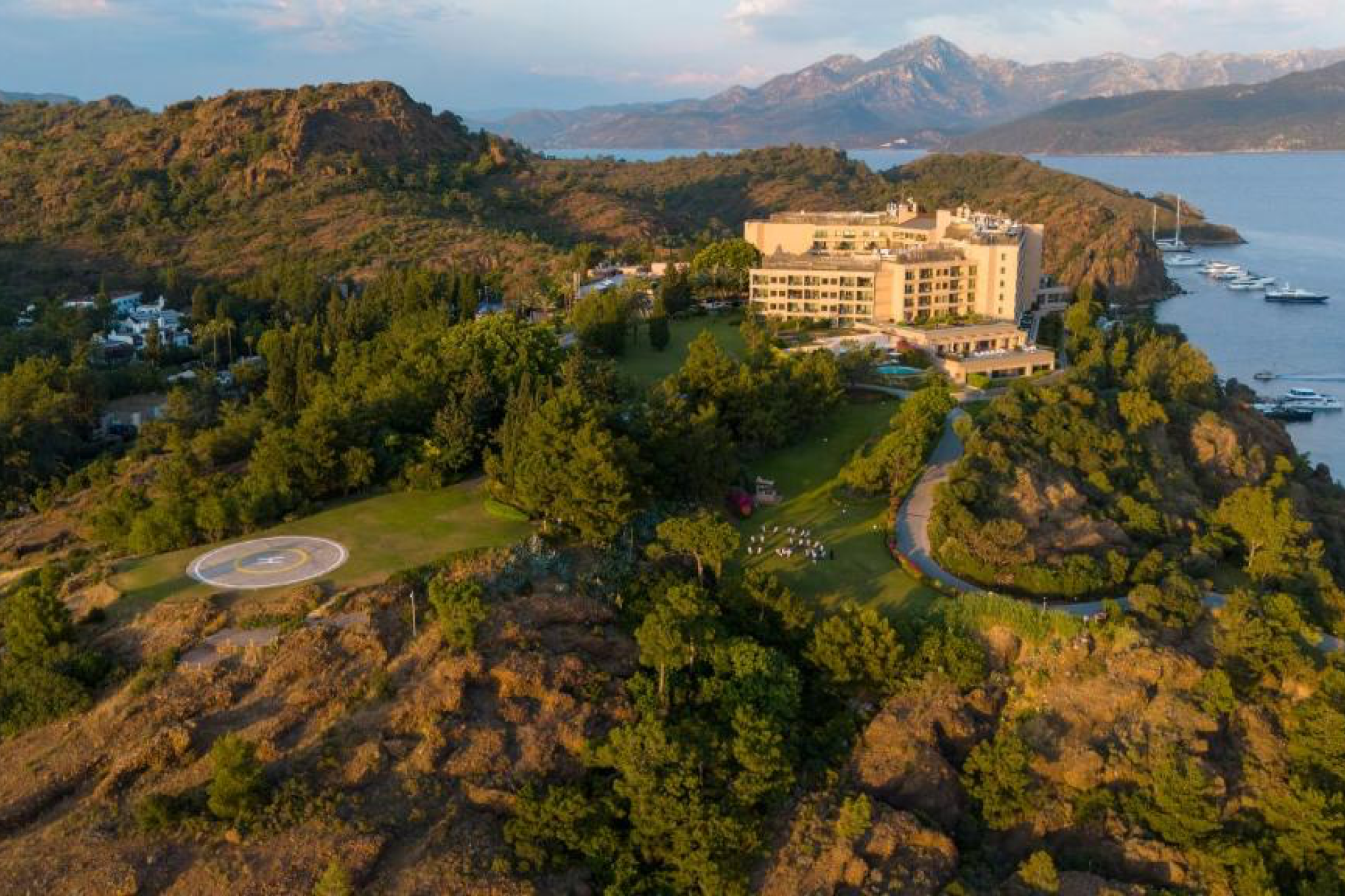 A sweeping aerial view of D Maris Bay perched above a secluded cove, surrounded by rugged mountains and yachts anchored in turquoise water—an exclusive retreat counted among the top beach resorts in Europe in 2025.