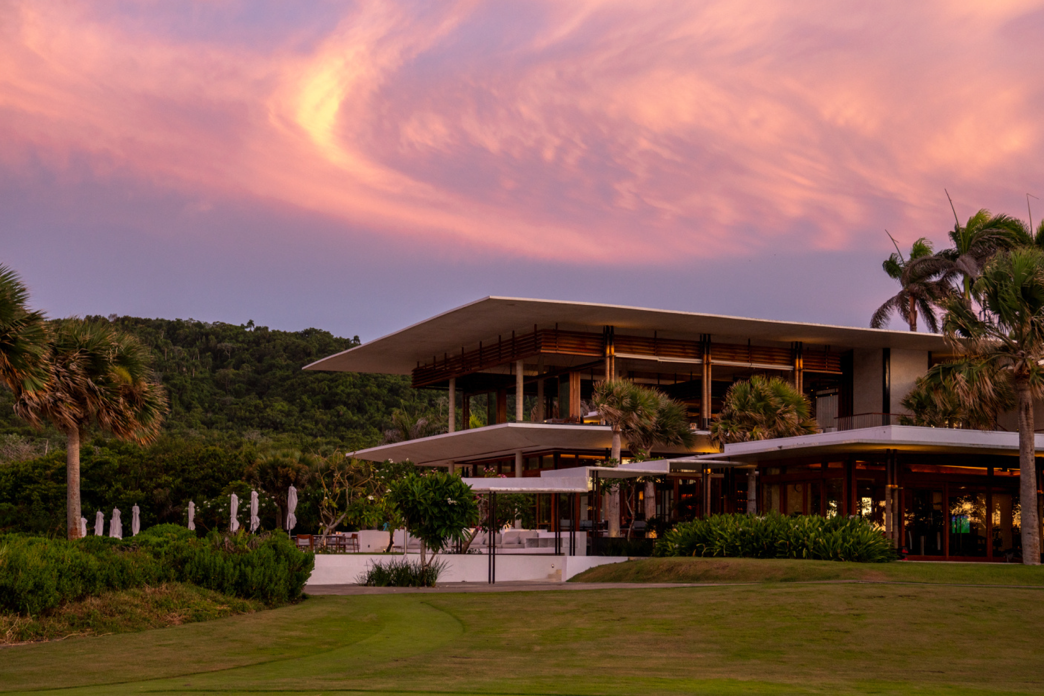 A dramatic sunset view of Amanera’s cliffside modernist pavilion framed by swaying palms and lush hills, glowing softly under swirling pink skies — a breathtaking architectural standout among the top resorts in the Caribbean Islands in 2025.