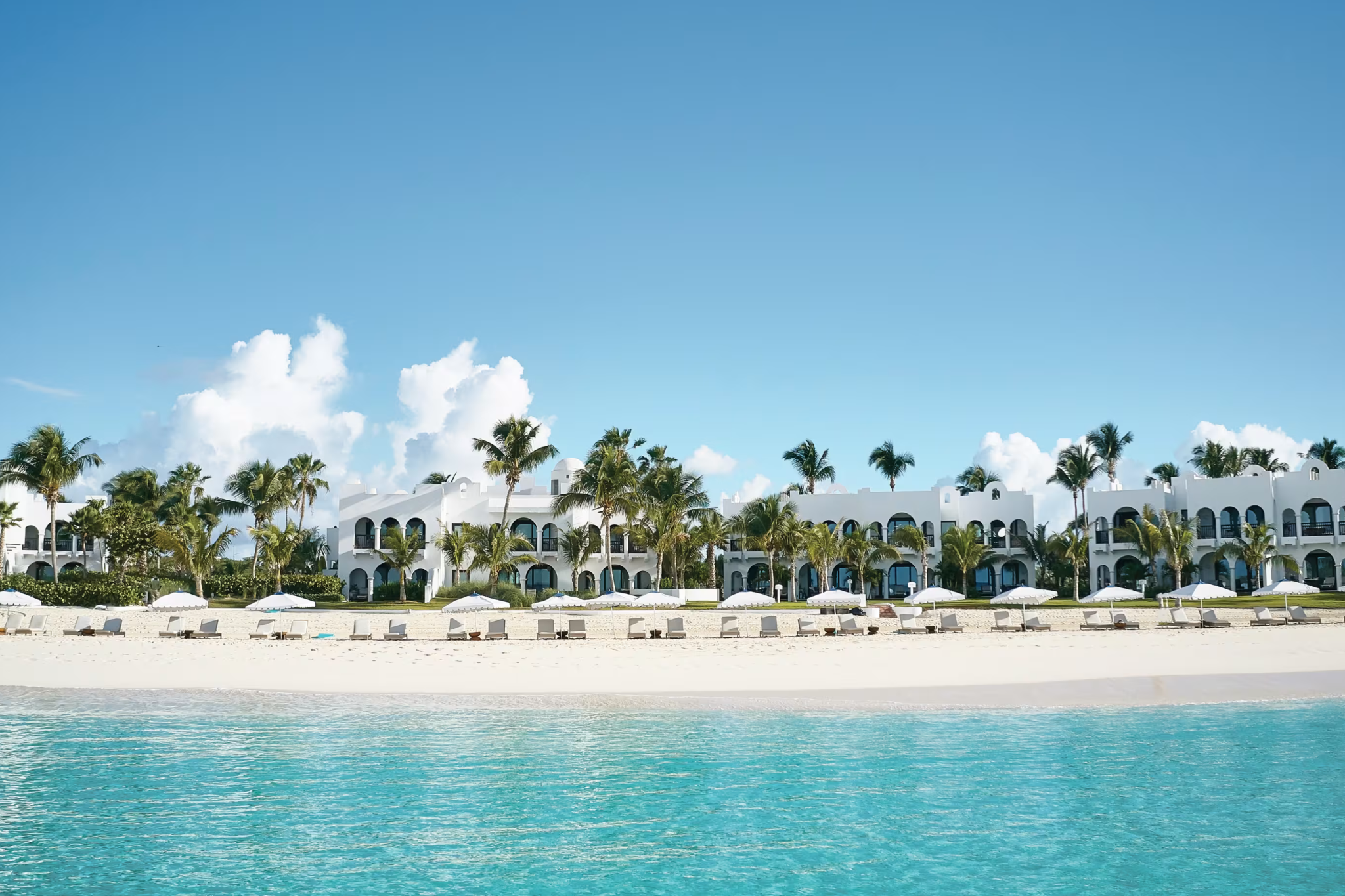 A panoramic beachfront view of Cap Juluca’s white Moorish-inspired architecture lined with palm trees and shaded loungers along Maundays Bay’s glowing turquoise water — a luminous coastal retreat ranking among the top resorts in the Caribbean Islands in 2025.