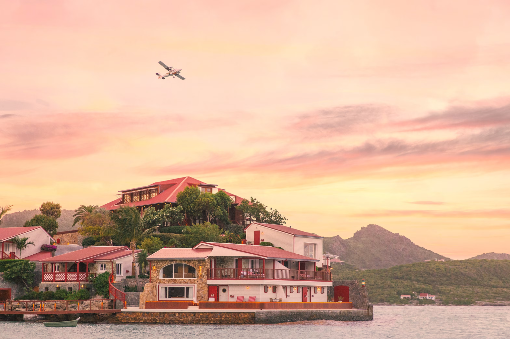 Eden Rock St Barths perched above the water at golden hour, with its red-roofed suites, rocky shoreline, and a seaplane flying overhead — a glamorous, iconic destination recognized as one of the top resorts in the Caribbean Islands in 2025.