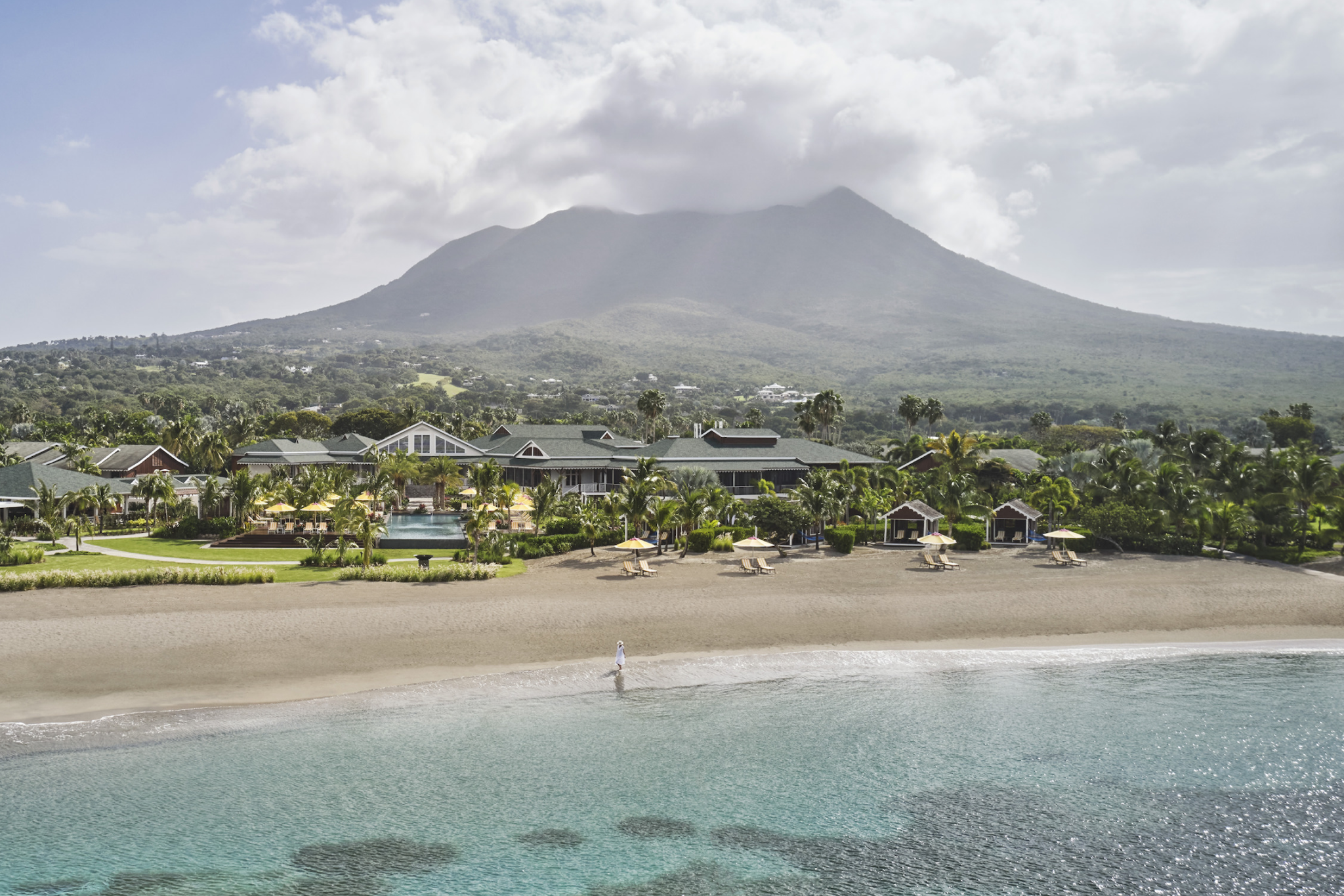 A wide beachfront view of Four Seasons Resort Nevis, showing golden sand, calm turquoise water, and lush tropical gardens at the base of Nevis Peak — a serene scene representing one of the top resorts in the Caribbean Islands in 2025.