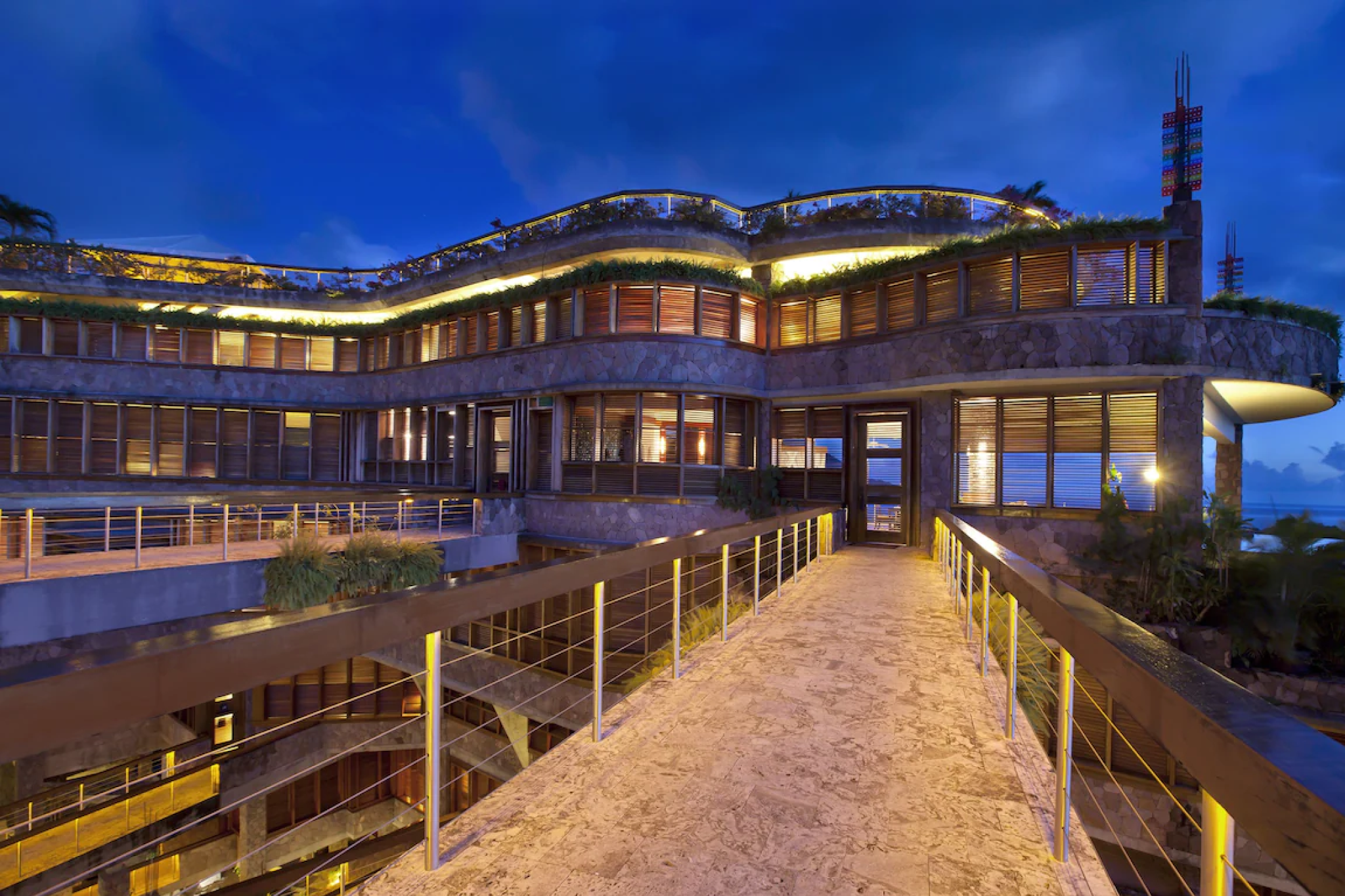 A nighttime exterior of Jade Mountain’s stone-and-wood architectural masterpiece, featuring open terraces and glowing ambient lighting connected by a dramatic walkway — a bold, design-driven sanctuary considered one of the top resorts in the Caribbean Islands in 2025.