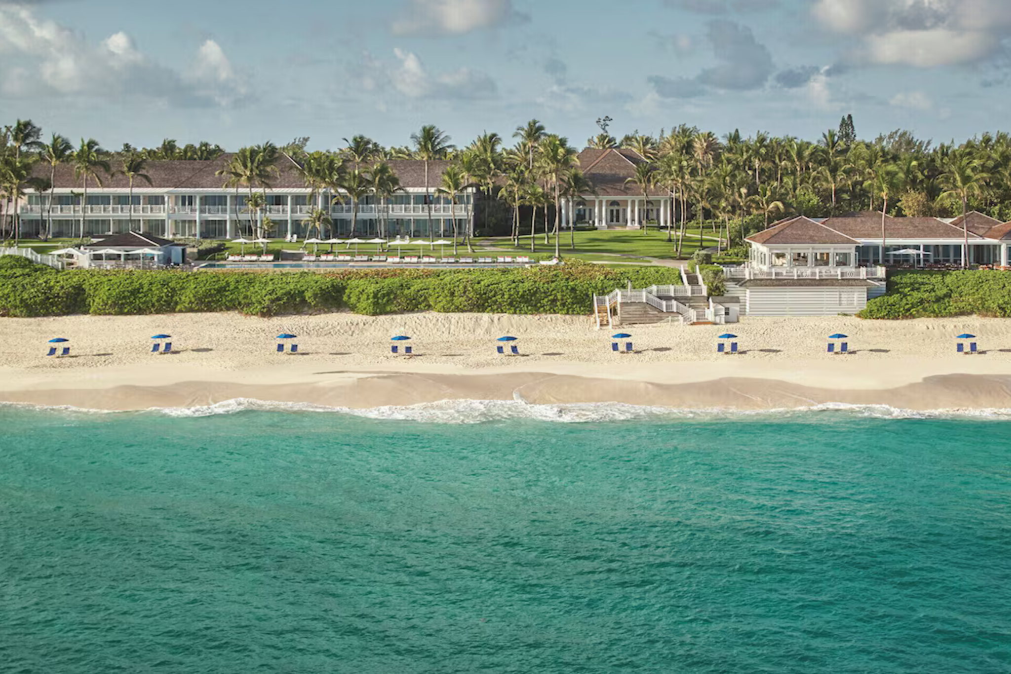 Aerial shoreline image of The Ocean Club in the Bahamas, featuring manicured lawns, white colonial-style buildings, and rows of blue-striped beach loungers along powdery white sand — a refined retreat celebrated as one of the top resorts in the Caribbean Islands in 2025.