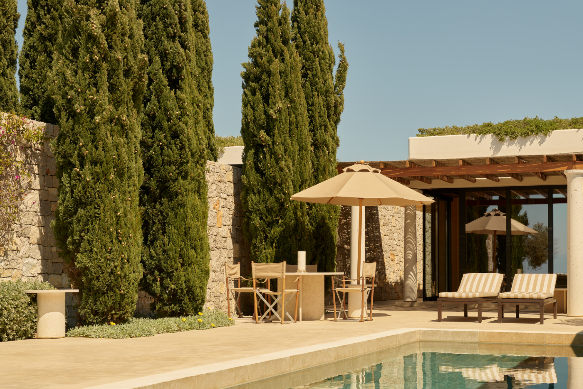 A serene courtyard scene at Amanzoe with stone walls, tall cypress trees, shaded loungers, and a tranquil pool reflecting the soft Mediterranean light — a minimalist, temple-inspired sanctuary leading the list of the top resorts in the Mediterranean in 2025.