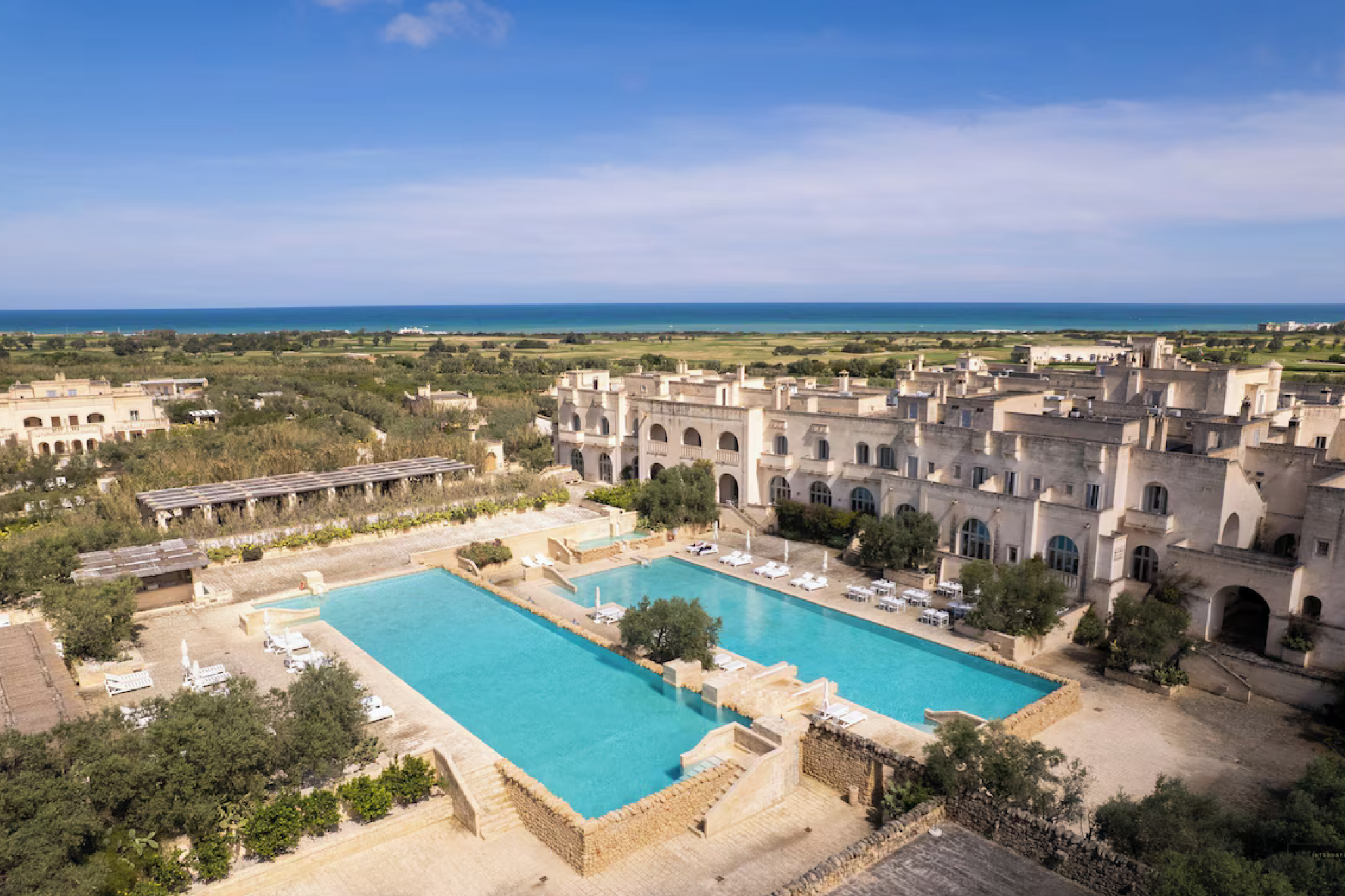 An expansive aerial view of Borgo Egnazia, showcasing limestone buildings, twin turquoise pools, olive groves, and distant Adriatic coastline — an immersive Puglian village-style property distinguished as one of the top resorts in the Mediterranean in 2025.