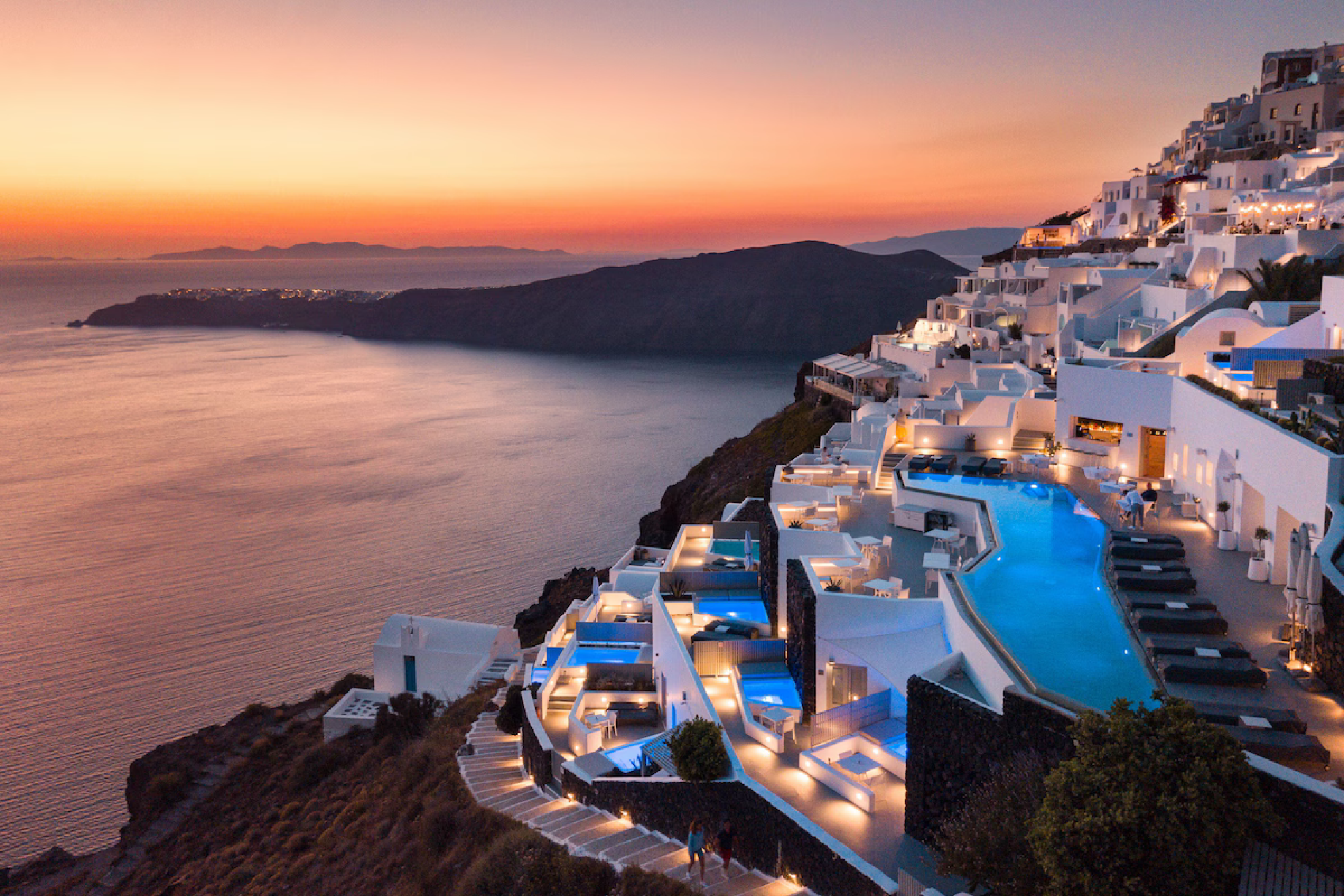 A dramatic sunset view of Grace Hotel in Santorini, with whitewashed cliffside suites, glowing infinity pools, and sweeping caldera vistas overlooking the Aegean Sea — a signature scene representing one of the top resorts in the Mediterranean in 2025.