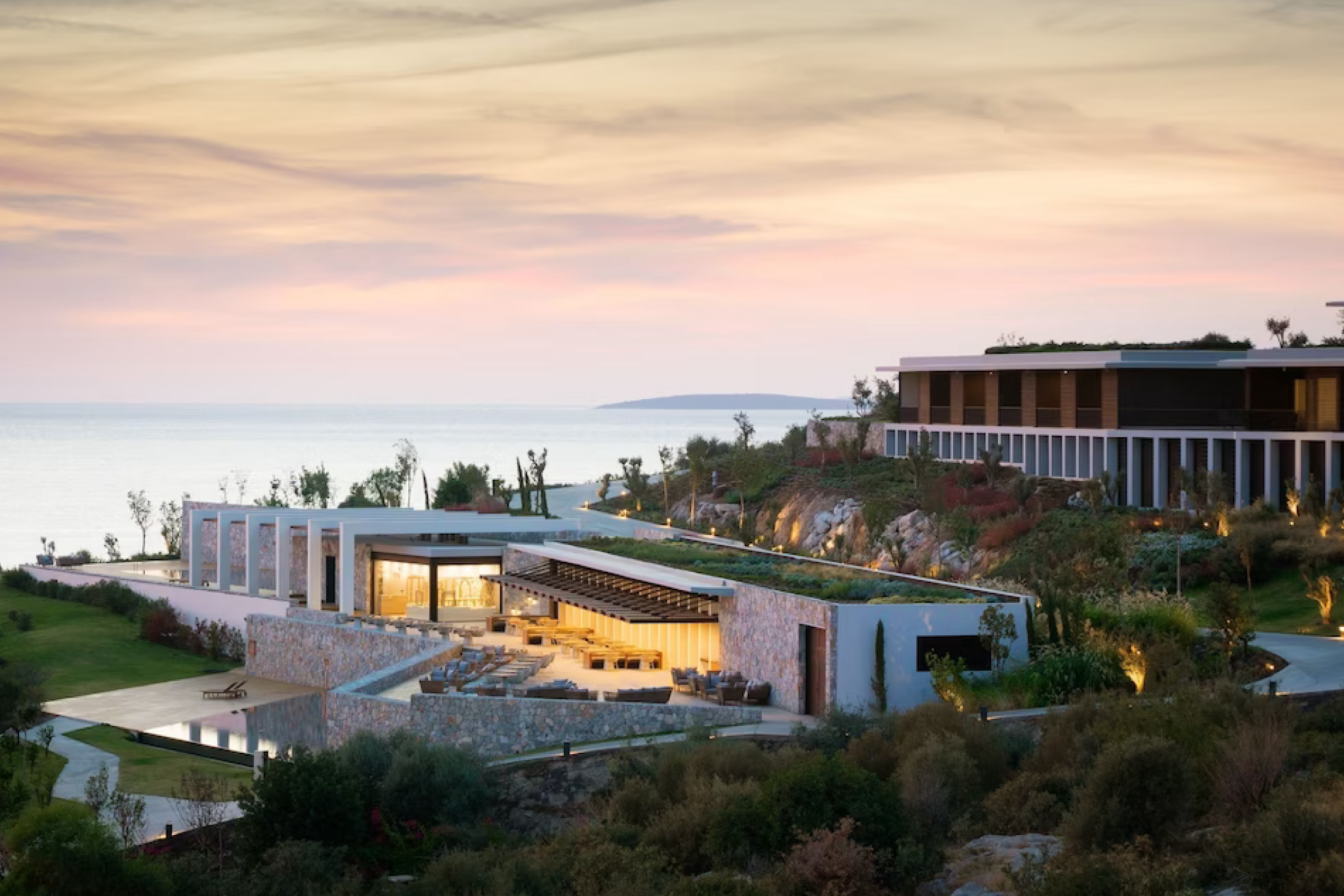 A panoramic hillside view of Six Senses Kaplankaya, featuring modern stone architecture, terraced structures, lush greenery, and tranquil Aegean waters at sunset — a wellness-driven hideaway celebrated as one of the top resorts in the Mediterranean in 2025.