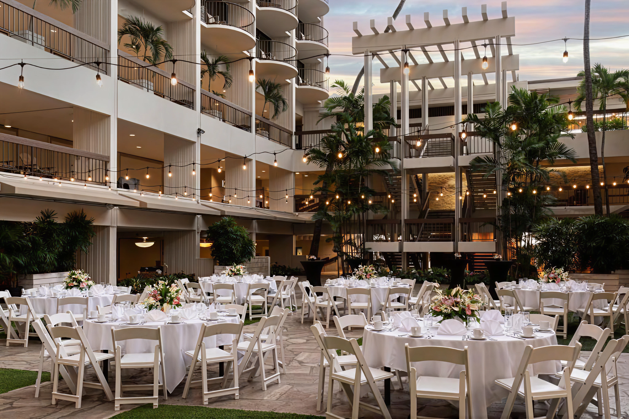 Courtyard event venue at waikiki beach marriott resort and spa arranged for a wedding or dinner, with round tables, white chairs, floral centerpieces, and overhead string lights.