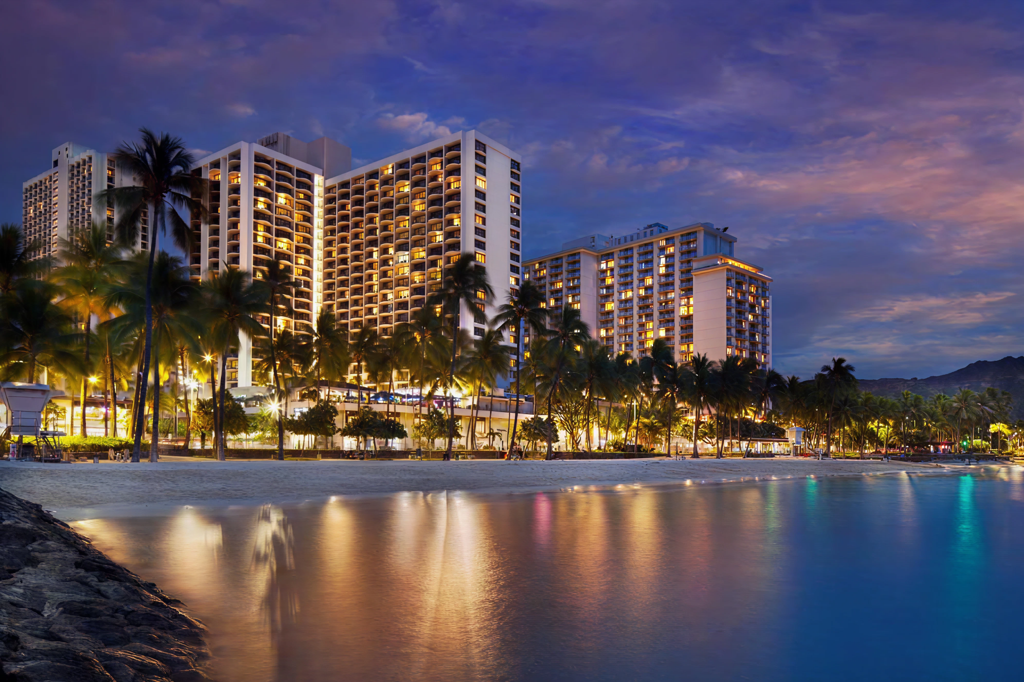 The oceanfront towers of waikiki beach marriott resort and spa lit up at twilight with palm trees lining the beach and reflections shimmering in the calm water of waikiki.