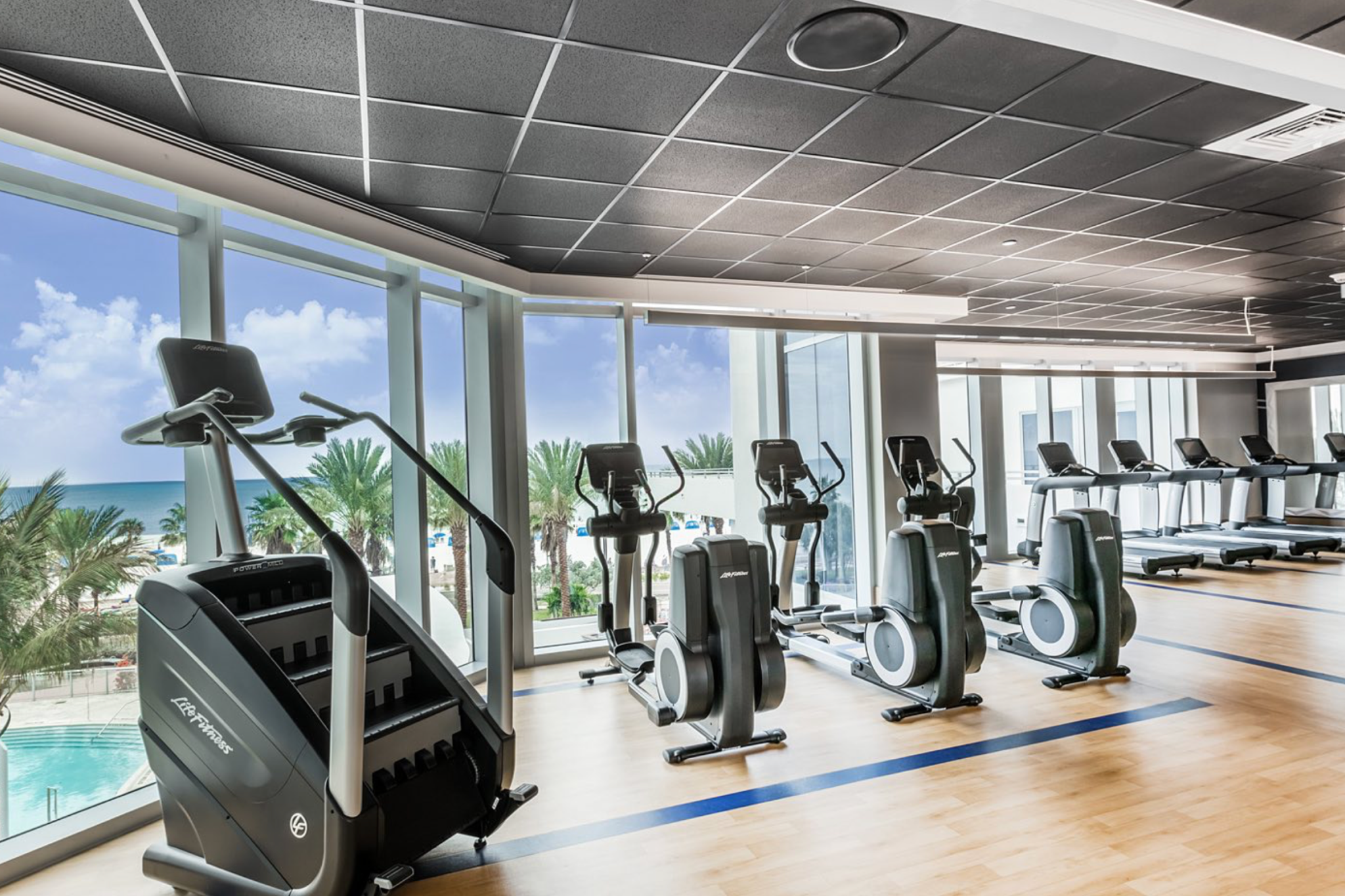 Oceanfront fitness center at Wyndham Grand Clearwater Beach with cardio equipment, floor-to-ceiling windows, and views of the pool and Gulf.